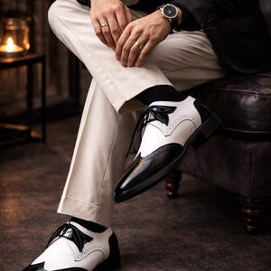 May include: Black and white spectator shoes with a classic design. The shoes feature a two-tone color scheme, with black leather and white accents. The shoes have a lace-up closure and a low heel. The shoes are part of a formal wear collection.