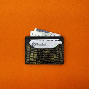 May include: A black cardholder with a textured, crocodile-like pattern in shades of green and brown. The wallet holds a driving license and currency. The background is a solid orange.