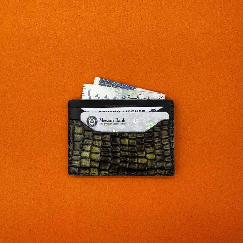 May include: A black cardholder with a textured, crocodile-like pattern in shades of green and brown. The wallet holds a driving license and currency. The background is a solid orange.
