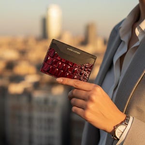 May include: A two-tone leather cardholder with a burgundy crocodile-embossed section and a black top. The cardholder is held up against a blurred cityscape background. The text "SLICK& LEATHER" is printed on the top.