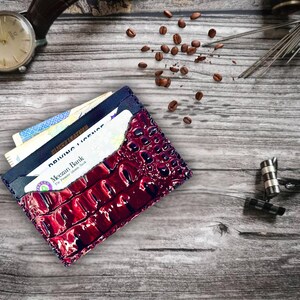 May include: A burgundy leather wallet with a crocodile skin texture, filled with cards and cash, sits on a wooden surface. A brown leather watch, coffee beans, and other accessories are also present.