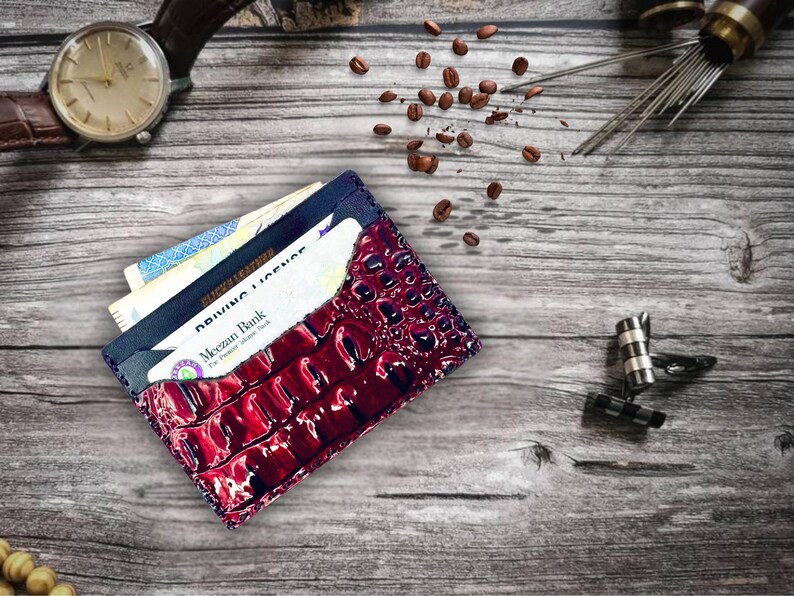 May include: A burgundy leather wallet with a crocodile skin texture, filled with cards and cash, sits on a wooden surface. A brown leather watch, coffee beans, and other accessories are also present.
