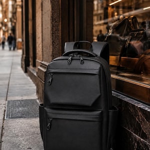 Könnte beinhalten: Ein schwarzer Rucksack mit mehreren Reißverschlussfächern und einem oberen Griff. Der Rucksack besteht aus einem glatten, möglicherweise lederartigen Material. Er steht auf einem Bürgersteig vor einem Geschäft.