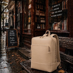 May include: A beige backpack with a front pocket and gold zippers sits on a wooden bench in front of a bookstore. The store's sign reads "The Book Nook." The window displays books and the quote, "A book is a dream that you hold in your hands."