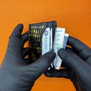 May include: A black leather wallet with a crocodile skin texture, holding a bank card, a driver's license, and several bills. The wallet is held by gloved hands against an orange background.