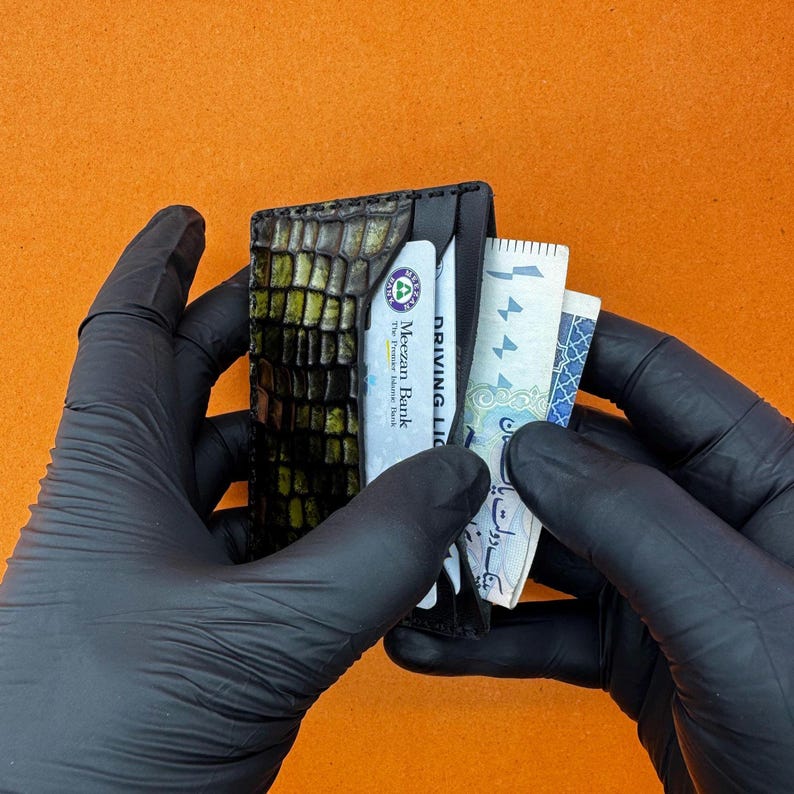 May include: A black leather wallet with a crocodile skin texture, holding a bank card, a driver's license, and several bills. The wallet is held by gloved hands against an orange background.