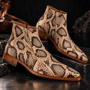 Könnte beinhalten: Ein Paar Stiefeletten mit einem beigen und dunkelbraunen Schlangenledermuster. Die Stiefel haben eine spitz zulaufende Zehenpartie, einen seitlichen Reißverschluss und einen gestapelten Holzabsatz mit einem kleinen dunklen Logo an der Seite.