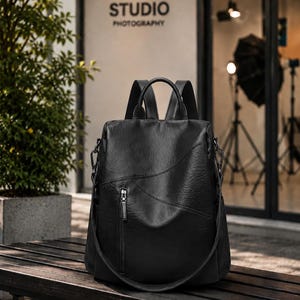 May include: Black leather backpack with a top handle, adjustable shoulder straps, and a front zippered pocket. The bag is sitting on a wooden bench in front of a building with the word "STUDIO PHOTOGRAPHY" on the wall.