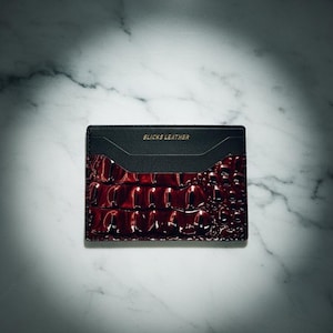 May include: A black and burgundy leather card holder with the text "SLICKS LEATHER" in gold lettering. The card holder has a textured, crocodile-like pattern on the burgundy section. The background is a white marble surface.