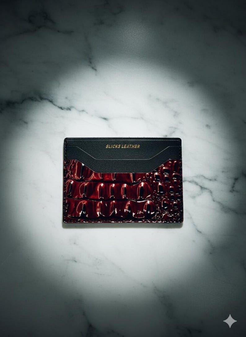 May include: A black and burgundy leather card holder with the text "SLICKS LEATHER" in gold lettering. The card holder has a textured, crocodile-like pattern on the burgundy section. The background is a white marble surface.