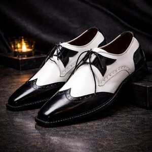 May include: A pair of two-tone dress shoes. The shoes are black and white, with a glossy finish. The toe and heel are black, while the upper is white. The shoes have a classic lace-up design and decorative perforations.