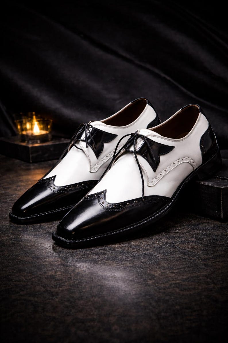 May include: A pair of two-tone dress shoes. The shoes are black and white, with a glossy finish. The toe and heel are black, while the upper is white. The shoes have a classic lace-up design and decorative perforations.