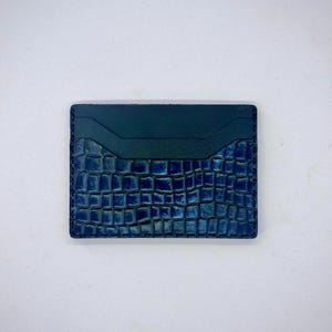 以下が含まれることがあります： クロコダイル柄の青い革製カードホルダー。上部は無地の濃い青色で、下部はテクスチャ加工されたエンボス加工のパターンです。カードホルダーには複数のカードスロットがあります。
