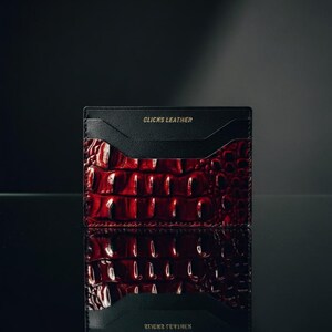 May include: A red and black leather card holder with a crocodile skin pattern. The card holder has the words "CLICKS LEATHER" printed in gold on the top and bottom. The card holder is displayed on a reflective surface.