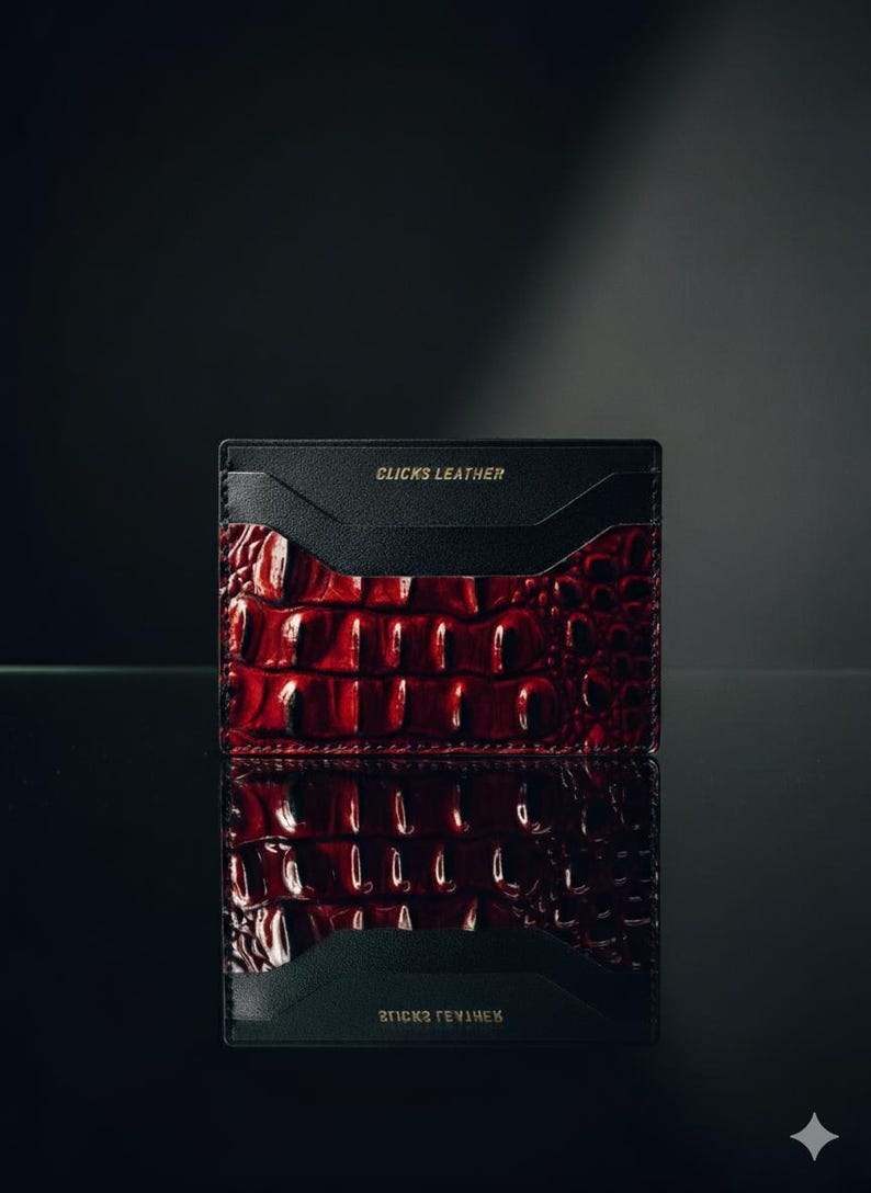 May include: A red and black leather card holder with a crocodile skin pattern. The card holder has the words "CLICKS LEATHER" printed in gold on the top and bottom. The card holder is displayed on a reflective surface.