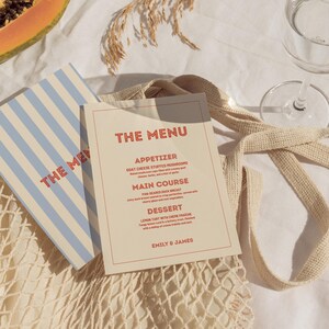 May include: Two menus with the text "THE MENU" in red. One menu has blue and white stripes. The other menu is beige with a red border and lists an appetizer, main course, and dessert. A glass of water and a net bag are also visible.