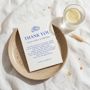 May include: A thank you card with blue text and a double ring graphic, resting on a speckled ceramic plate. A glass of water with a lemon slice and a wheat stalk are nearby. The card reads "THANK YOU" and a message.