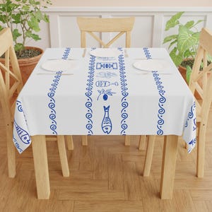Peut inclure: Une nappe blanche ornée d'une bordure bleue en forme de clé grecque et d'un motif central représentant des poissons, des olives et des verres à vin. La nappe est posée sur une table en bois avec des chaises assorties.
