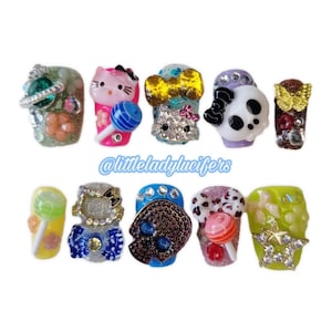 May include: A collection of ten press-on nails with various designs. The nails feature glitter, rhinestones, and charms, including a Hello Kitty, a skull, and a butterfly. The nails are in various colors, including pink, blue, and green. The text "@littleladyglucifers" is visible.