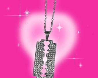 May include: Silver-toned pendant necklace featuring a razor blade design, encrusted with small, sparkling crystals. The pendant hangs from a delicate chain. The background is a gradient of pink and white with star-shaped sparkles.