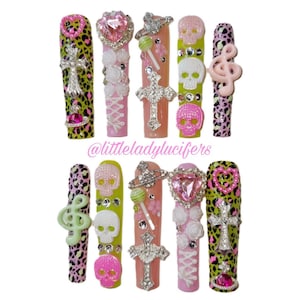 Op de afbeelding: Een set van tien lange kunstnagels met diverse ontwerpen, waaronder groene en roze luipaardprints, zilveren kruisbedels, roze hartvormige edelstenen, witte schedelapplicaties, lollybedels en muzikale vioolsleutels met de tekst @littleladylucifers.