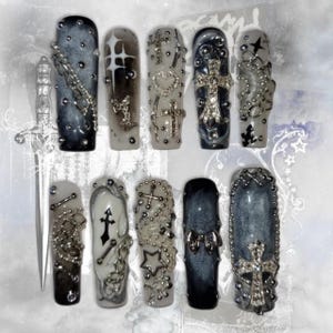 Puede incluir: Un conjunto de diez uñas artificiales largas en forma de ataúd. Cada uña presenta un diseño único con adornos plateados, incluyendo cruces, cadenas y estrellas, sobre un degradado de negro, gris y blanco. También hay una espada decorativa.
