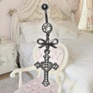 May include: A silver-toned belly button ring with a dangling cross pendant. The top ball and the center jewel are clear. A bow-shaped charm connects the jewel to the cross. The cross is encrusted with small, clear stones.