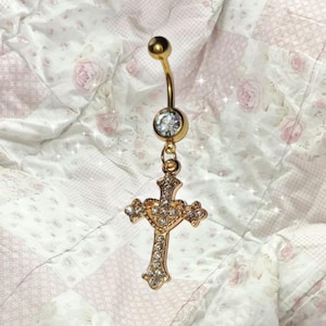 May include: Gold-coloured belly button ring with a curved barbell design. The top ball and the bottom charm are adorned with clear crystals. The charm is a detailed cross shape, also embellished with crystals.