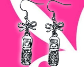 May include: Silver-toned dangle earrings featuring a bow and a flip phone charm. The earrings have a hook closure and are set against a pink and white background. The flip phone charm has a detailed design.