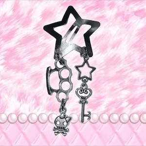 May include: Silver hair clip with a star-shaped barrette. The clip has charms hanging from it, including a set of brass knuckles, a star, a key, and a skull with crossbones. The background is pink with a pearl border.