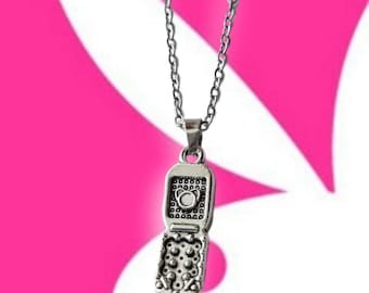 May include: A silver-colored pendant necklace featuring a detailed charm shaped like a flip phone. The phone charm is open, revealing the keypad and screen details. The necklace has a delicate chain and is set against a pink and white background.