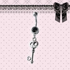 以下が含まれることがあります： 黒い宝石とスカルキーチャームが付いたシルバーのベリーボタンリング。キーにはハート型のディテールがあります。ジュエリーはピンクのドット柄の背景に表示されています。