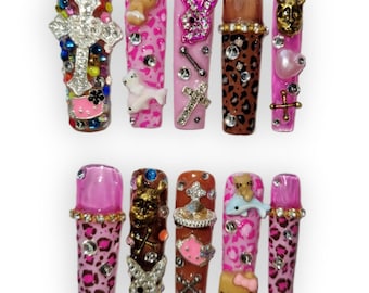 May include: A set of ten long, embellished artificial nails. Designs include crosses, a bunny, a heart, and a Hello Kitty. Colors include pink, brown, and leopard print, with rhinestones and other decorative elements.