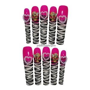 May include: A set of long, artificial nails with a vibrant pink base and zebra print design. Each nail is adorned with a pearl-lined heart or a gold skull with rhinestones. The nails are designed for a fashionable look.