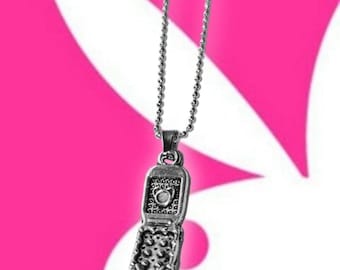May include: Silver-toned pendant necklace featuring a detailed, open flip phone charm. The charm has a textured surface with a heart design. The necklace hangs against a pink and white background.