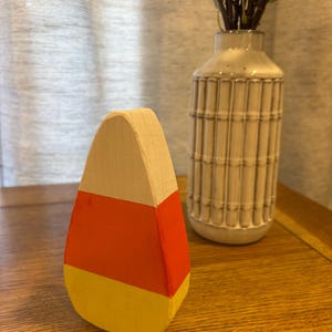 May include: A wooden candy corn decoration featuring a triangular shape with three horizontal color bands. The top section is white, the middle section is orange, and the bottom section is yellow. The surface has a matte, painted finish.