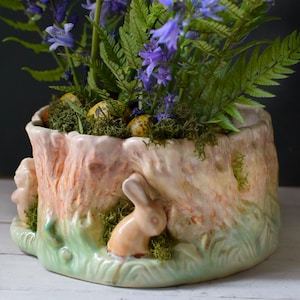 May include: A ceramic planter shaped like a tree stump, filled with bluebell flowers, ferns, and speckled eggs. The planter has a light pink and green colour scheme with two small bunny figurines on the side.