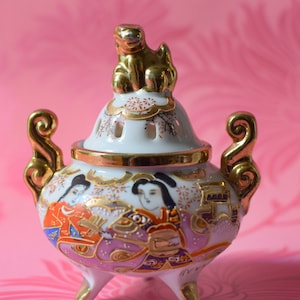 May include: A white porcelain incense burner with gold accents and a gold animal figurine on top. The burner features a floral design and three legs. The burner is decorated with painted figures of women in traditional Japanese clothing.