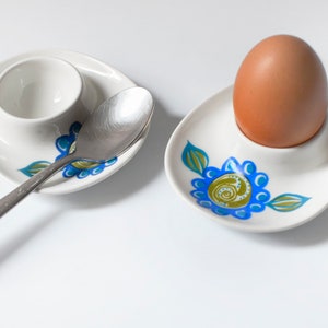 May include: Two white ceramic egg cups with blue and green floral designs. One egg cup has a brown egg inside. The other egg cup has a silver spoon next to it.