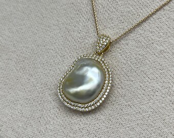 May include: A gold necklace featuring a large, irregularly shaped pearl pendant. The pearl is surrounded by a double halo of small, sparkling stones. The necklace chain is delicate and gold-toned, creating an elegant jewelry piece.