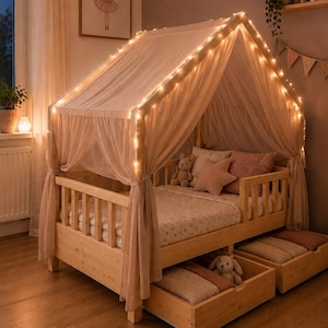 May include: A light-colored wooden house-shaped bed frame with a peaked roof draped in sheer fabric and illuminated by a string of warm white fairy lights. The bed includes two pull-out storage drawers underneath and features plush pillows.