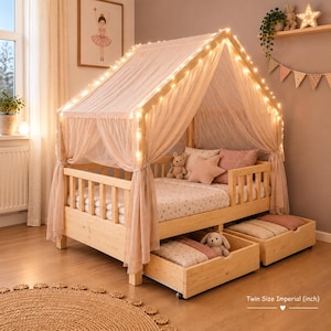 May include: A light-colored wooden house-shaped bed frame with a twin size imperial mattress, draped in sheer white fabric and illuminated by string lights. The bed features two pull-out storage drawers underneath and includes plush pillows.