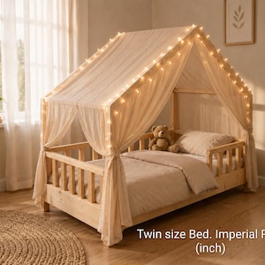 May include: A light-colored wooden house-shaped twin size bed frame with a peaked roof structure. The frame is draped with sheer beige fabric and illuminated by a string of warm white fairy lights. A teddy bear sits on the beige bedding.