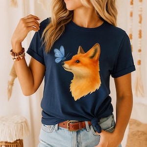 Può includere: T-shirt blu navy con un'illustrazione di una volpe arancione e una farfalla blu. Il design della volpe è dettagliato e accattivante. La maglietta è abbinata a pantaloncini di jeans e una cintura marrone, creando un look casual ispirato alla natura.