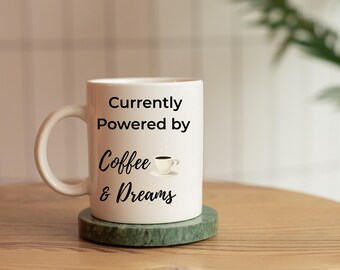 以下が含まれることがあります： 白いセラミック製のコーヒーカップで、黒い文字で「Currently Powered by Coffee & Dreams」と書かれています。コーヒーの文字の上に、コーヒーカップと蒸気を描いた小さな白黒のイラストがあります。カップは緑の大理石のコースターの上に置かれています。