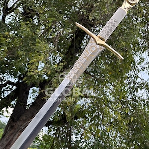 Peut inclure: Une longue épée avec une lame argentée et une poignée blanche. La poignée est ornée d'accents dorés et de motifs complexes. La garde et le pommeau sont également dorés, avec un motif en forme de soleil sur le pommeau. L'épée est posée sur un fond de feuillage vert.