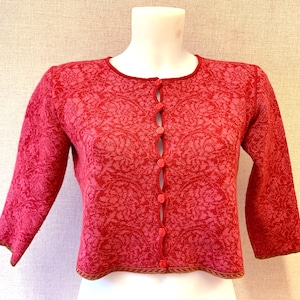 Pode incluir: Um cardigan rosa com um padrão floral e fecho de botões. O cardigan tem um comprimento curto e mangas três quartos.