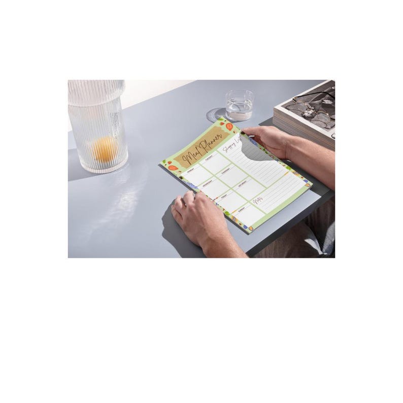 Puede incluir: Una persona sostiene un bloc de notas Meal Planner con un borde verde y una fuente manuscrita. El bloc de notas tiene secciones para la planificaci&oacute;n de comidas y una lista de compras. El fondo incluye un vaso de agua y un vaso decorativo.