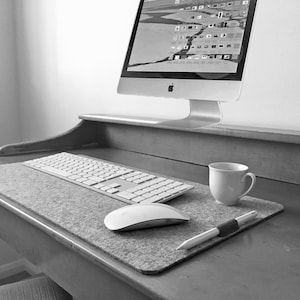 Puede incluir: Una alfombrilla de escritorio de fieltro gris con un teclado blanco, un ratón blanco y un bolígrafo plateado sobre un escritorio de madera. Una taza de café blanca está sobre la alfombrilla.
