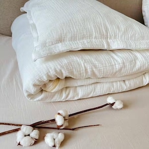 May include: A white cotton quilt folded on a bed with a white cotton pillow on top. The quilt is textured and has a subtle sheen. A few sprigs of cotton are in the foreground.